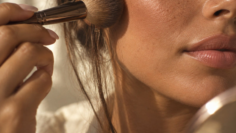 Close-up of a woman sweeping blush onto her cheek with a soft brush in warm morning light.