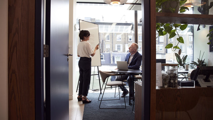 Woman presenting to colleagues at a wooden table in a warm plant-filled office