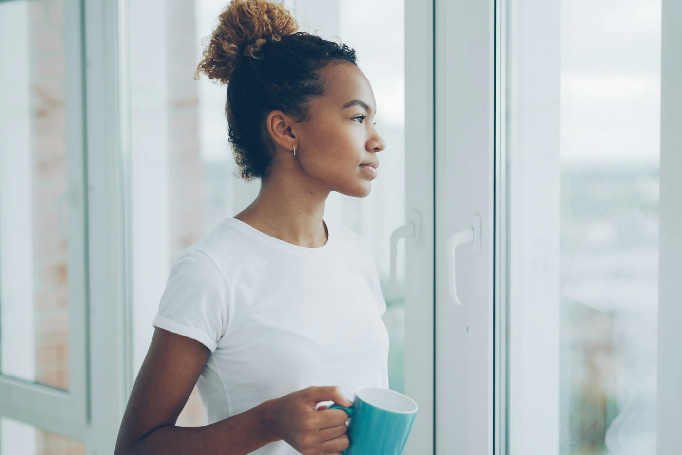 A woman holding a mug and looking out the window in warm, calm morning light at home